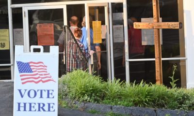 DOJ election monitors 2026 story: voters enter a polling place beside a prominent “Vote Here” sign.