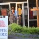 DOJ election monitors 2026 story: voters enter a polling place beside a prominent “Vote Here” sign.