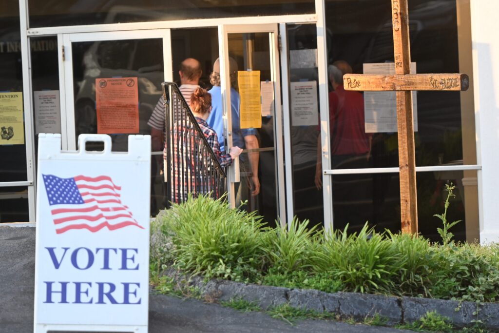 DOJ election monitors 2026 story: voters enter a polling place beside a prominent “Vote Here” sign.