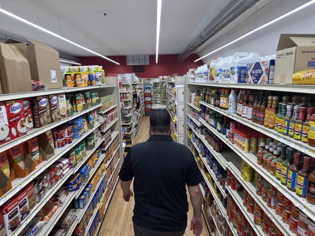 A shopper walks down a grocery aisle lined with shelves of packaged food, illustrating the Rising Cost of Living Midterms.