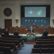 Lawmakers discussing the National Patient Safety Board Act of 2026 in a legislative chamber with digital graphics.
