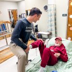 A caregiver helps an older man in a red outfit move from a bed in a care facility, illu — Cost of Caring for Aging Parents