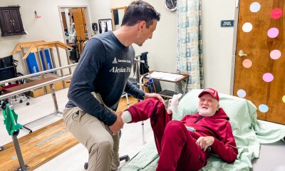 A caregiver helps an older man in a red outfit move from a bed in a care facility, illu — Cost of Caring for Aging Parents