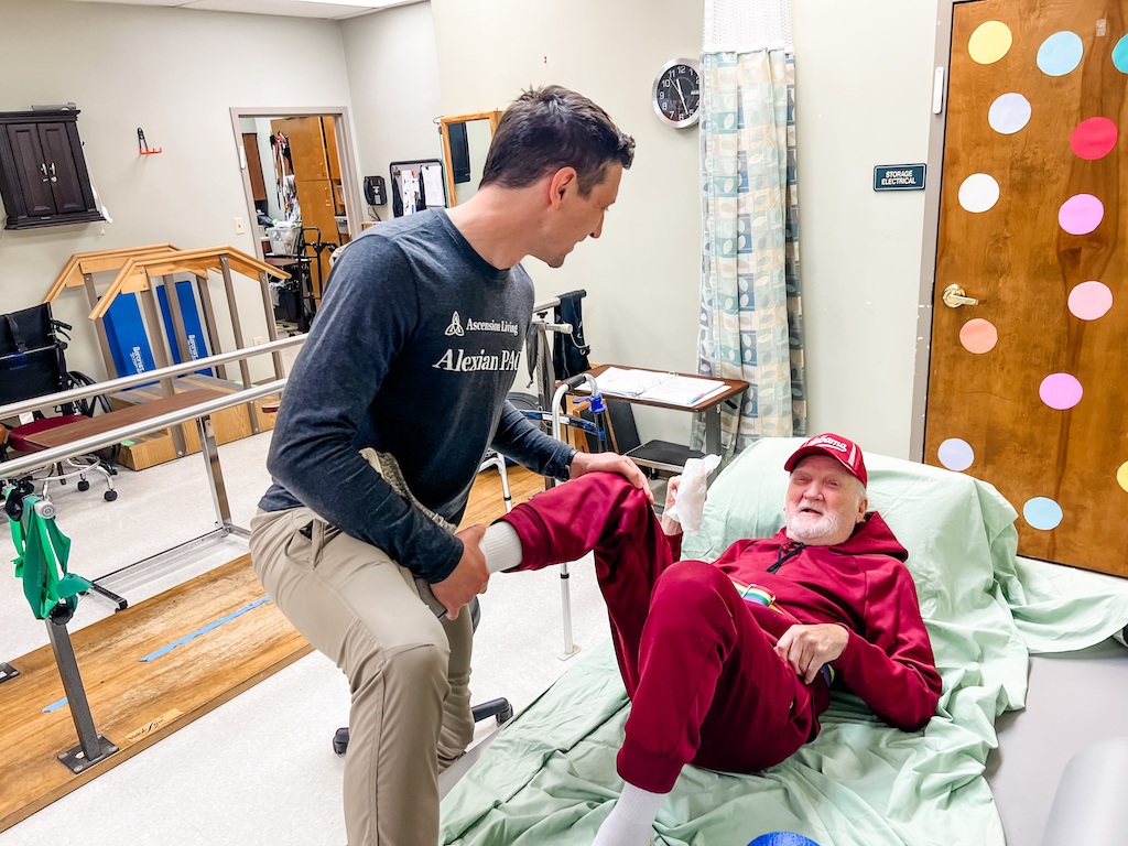 A caregiver helps an older man in a red outfit move from a bed in a care facility, illu — Cost of Caring for Aging Parents