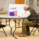 A voter in a green vest sitting at a table in a polling place, marking a ballot behind an — state laws on election betting
