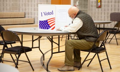 A voter in a green vest sitting at a table in a polling place, marking a ballot behind an — state laws on election betting