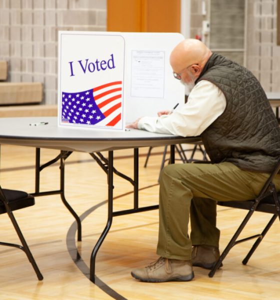 A voter in a green vest sitting at a table in a polling place, marking a ballot behind an — state laws on election betting