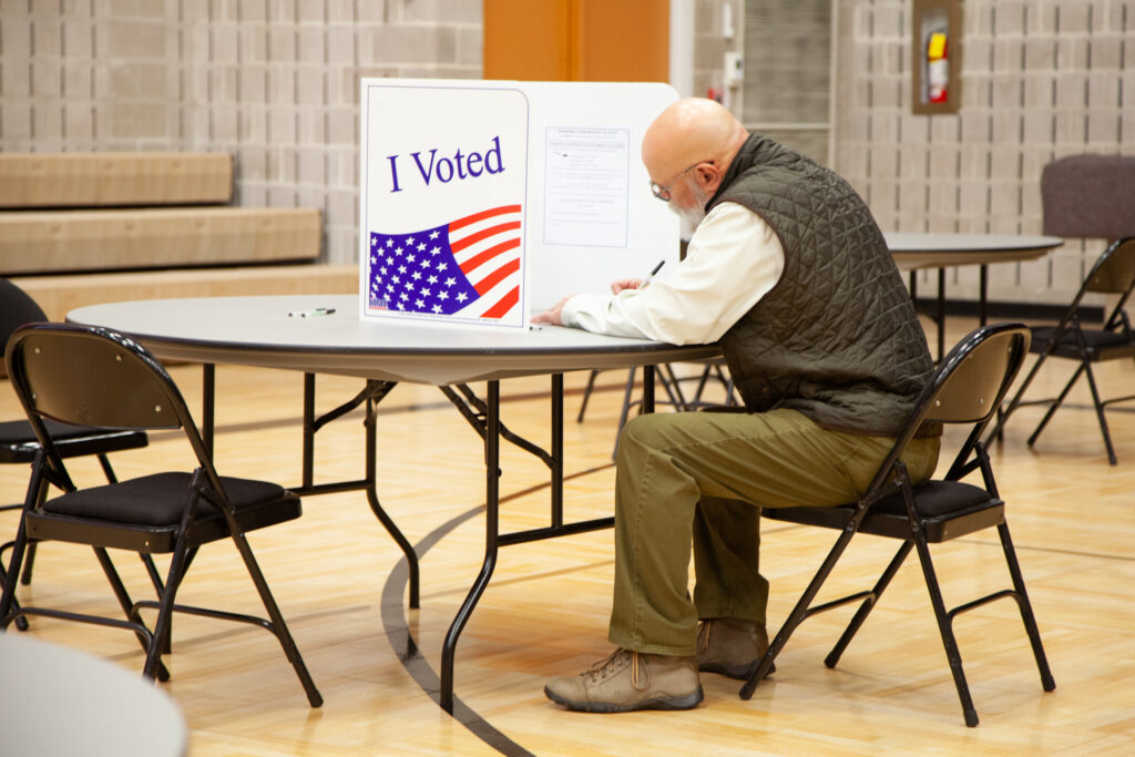A voter in a green vest sitting at a table in a polling place, marking a ballot behind an — state laws on election betting